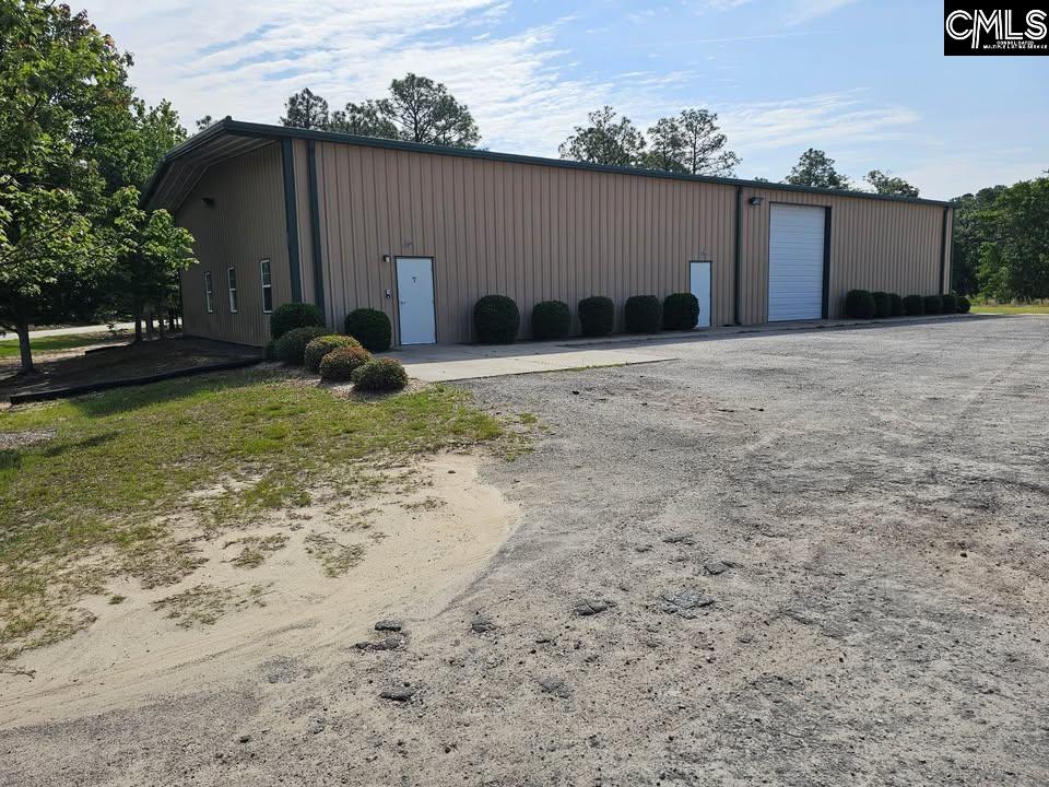 Tan metal commercial building with a large roll-up door beside a broad gravel parking area.