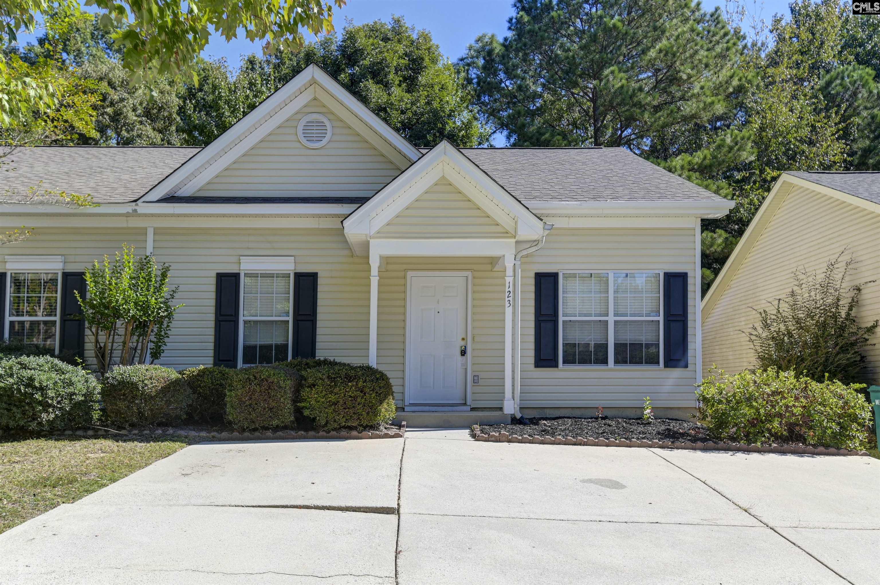 Beige single-story house with dark shutters, a covered entry, and a wide concrete driveway.