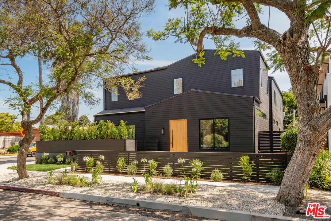 Modern dark-sided two-story home with a wood front door, black fence, and mature street trees.