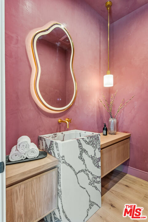 Pink bathroom with a veined marble basin, wood cabinetry, gold fixtures, and an irregular mirror.