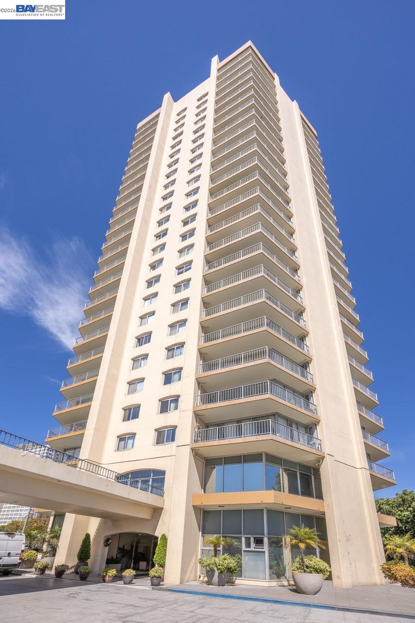 Beige residential tower with stacked balconies and a landscaped entrance beneath a clear blue sky.