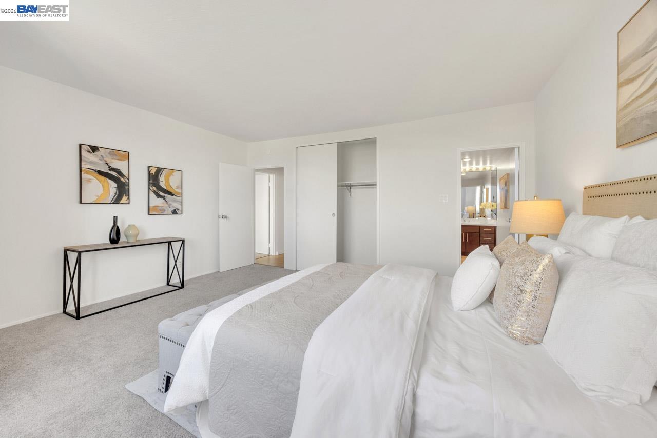 Staged bedroom with a large white bed, gray carpet, abstract art, and an open closet.