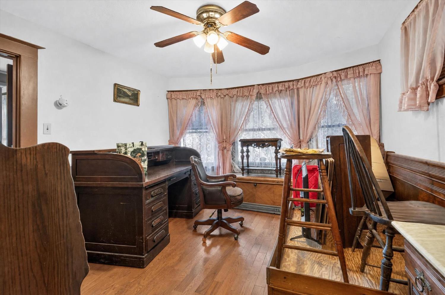 Home office with a dark wood desk, wooden chairs, hardwood floors, and pink curtains.