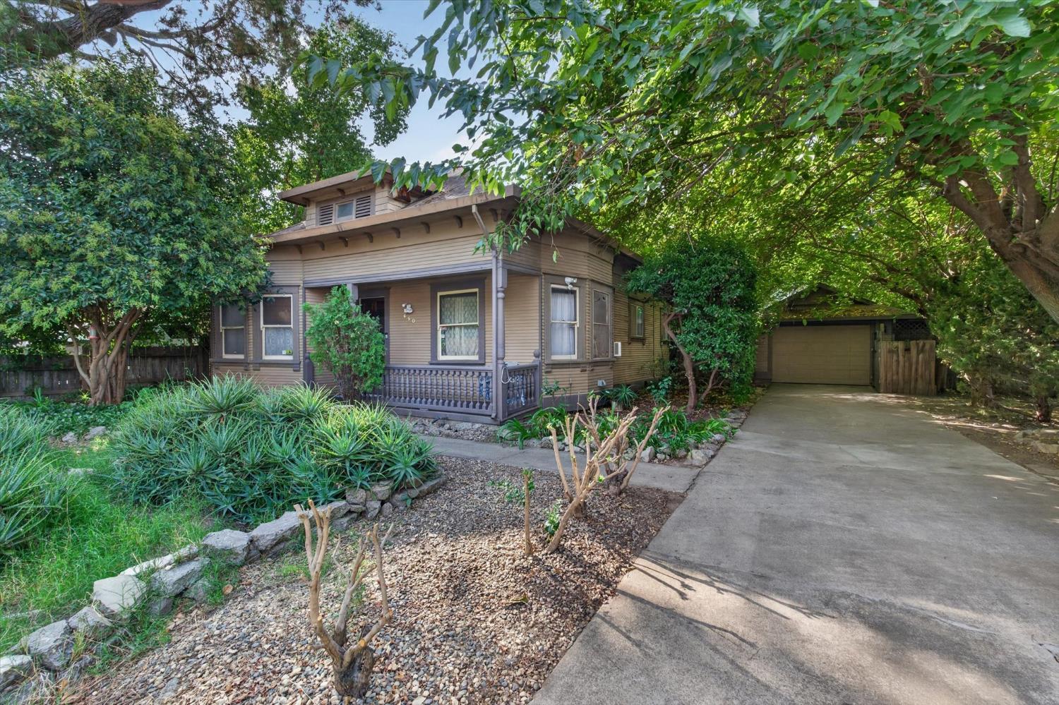 Two-story tan house with a covered porch, leafy trees, and a driveway leading to a detached garage.