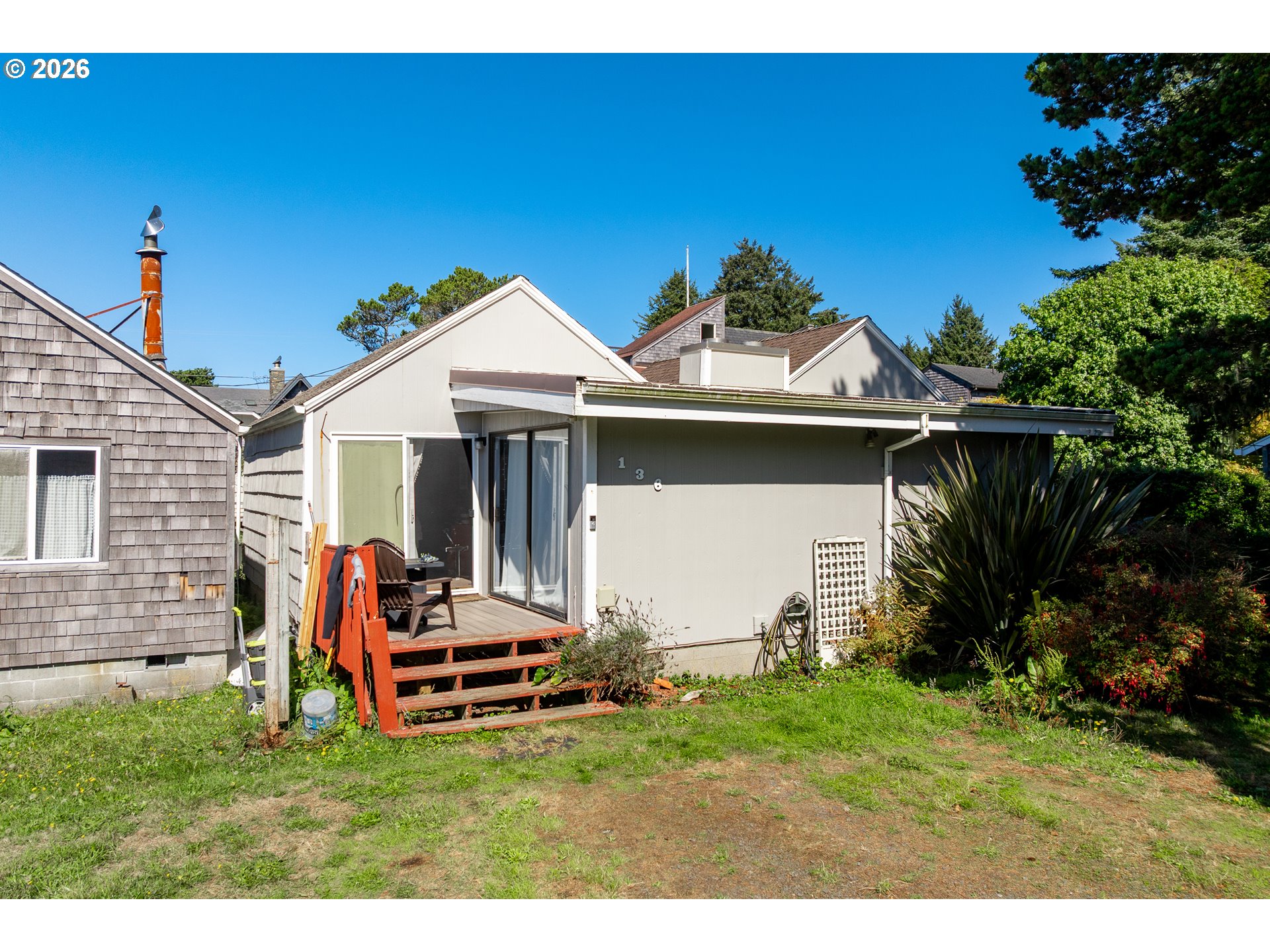 Coastal cottage exterior with a small red-stained deck, sliding doors, and surrounding garden.