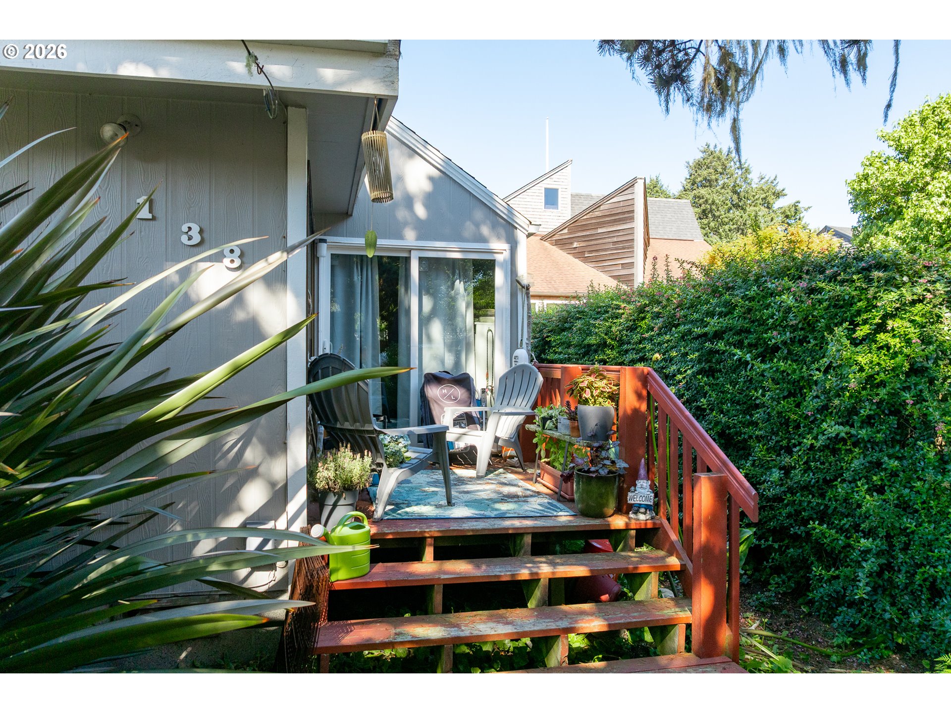 Home deck with Adirondack chairs, potted plants, and red-brown stairs beside a tall hedge.
