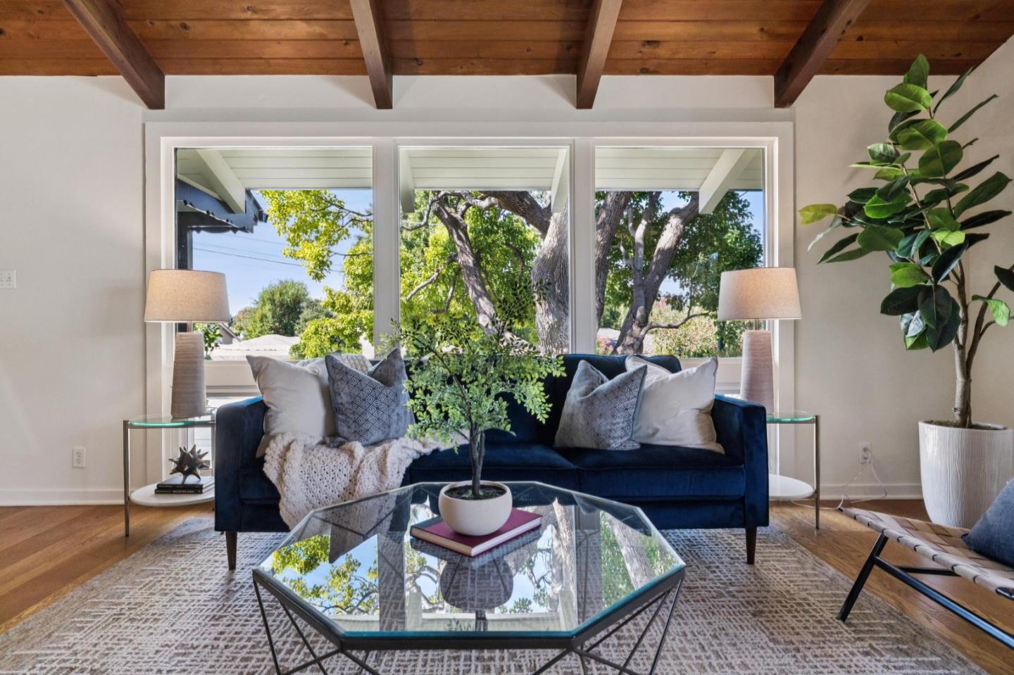 Living room with a navy sofa, glass coffee table, exposed beams, and large windows overlooking trees.