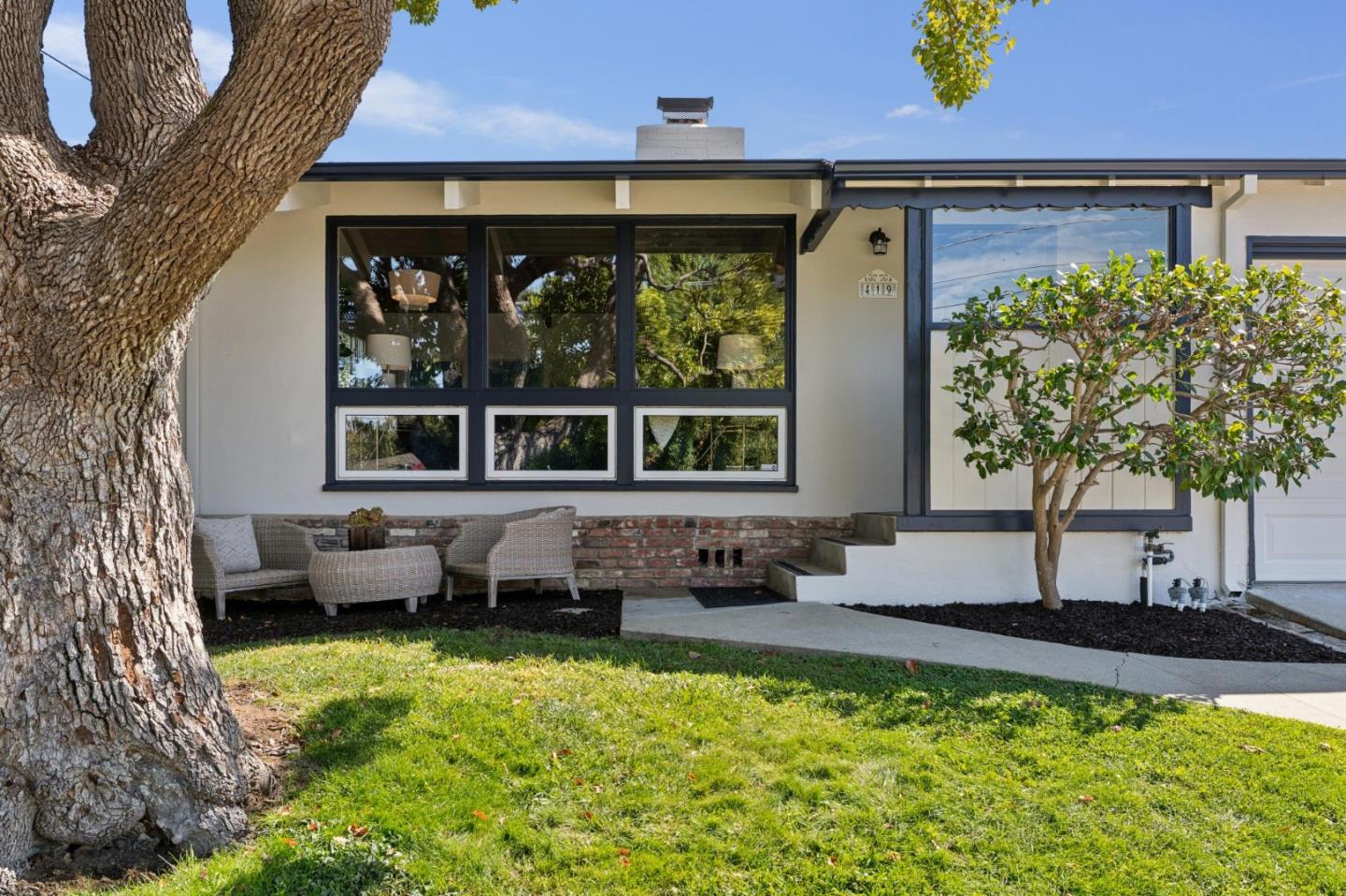 Single-story home with broad dark-framed windows, patio seating, and a large tree in the front lawn.