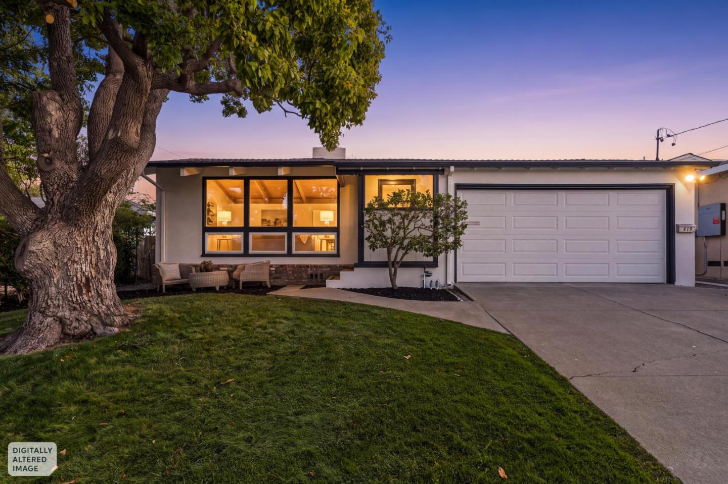 Single-story home with a large shade tree, glowing front windows, and an attached garage at dusk.
