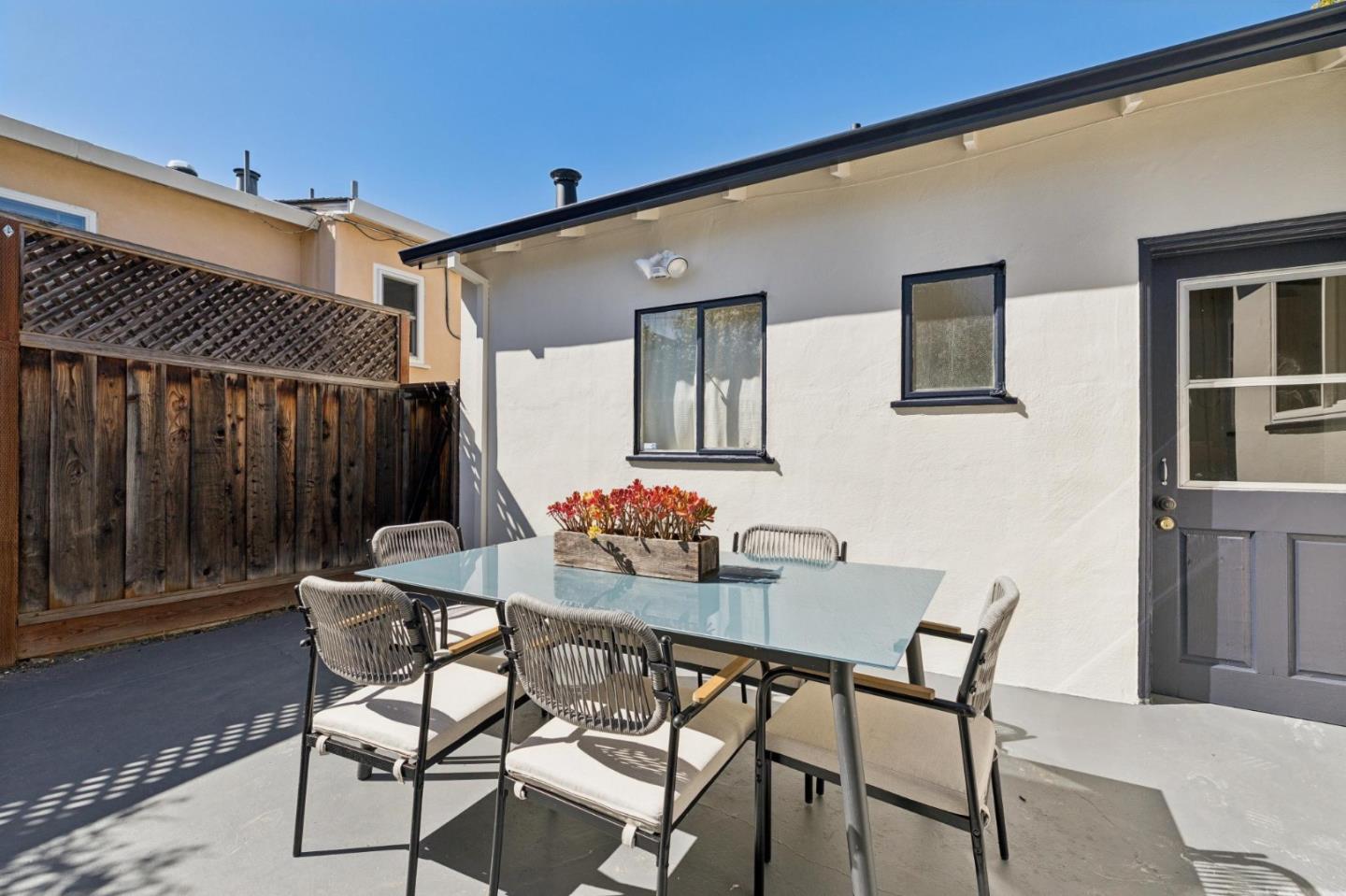 Back patio with a glass-top dining table and cushioned chairs beside a white stucco wall.