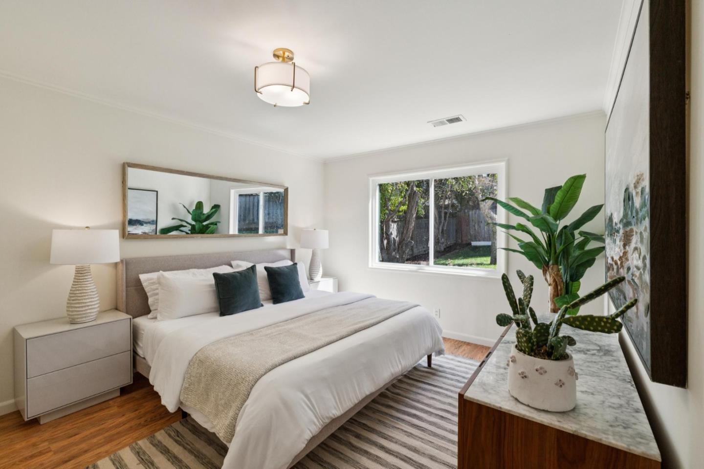 Staged bedroom with a large white bed, teal pillows, and a window overlooking a tree-lined yard.