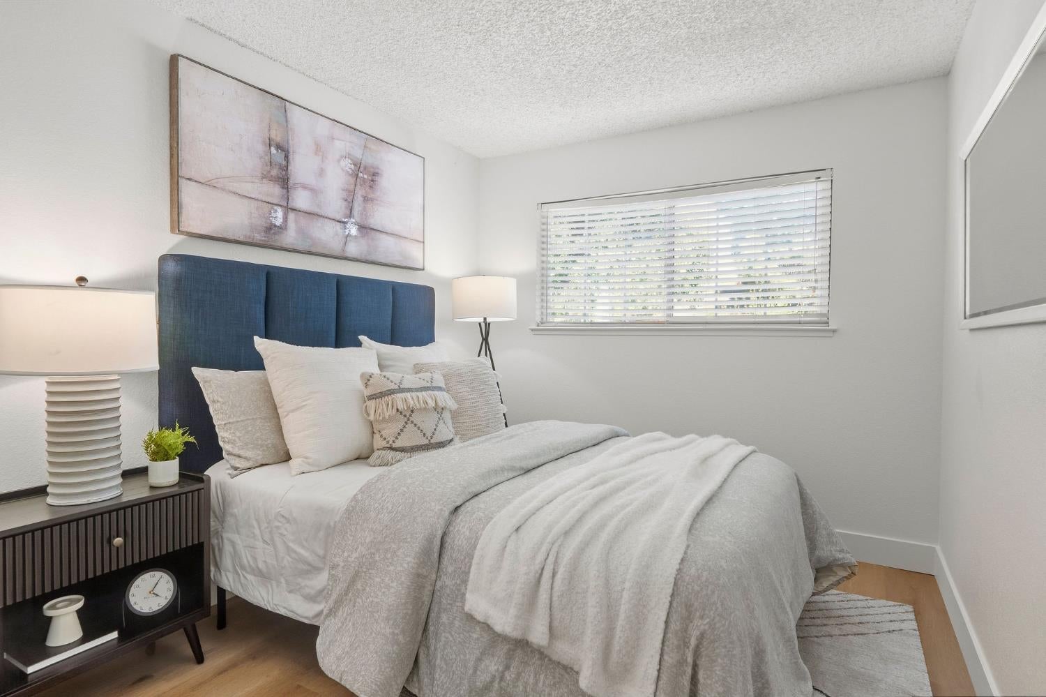 Bedroom with a blue upholstered headboard, layered neutral bedding, and a window with blinds.