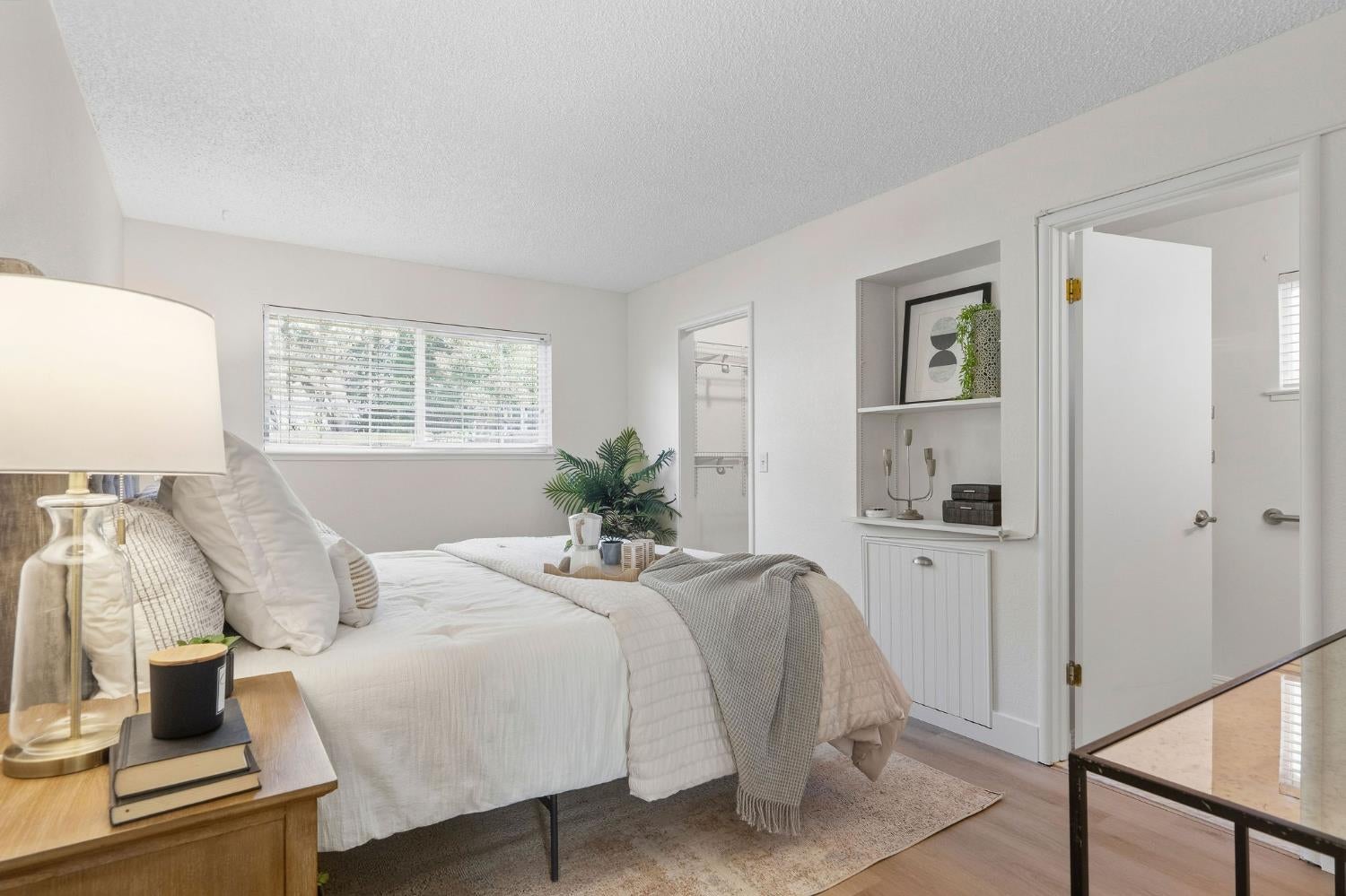 Staged bedroom with a white bed, neutral bedding, built-in shelves, and a wide window with blinds.