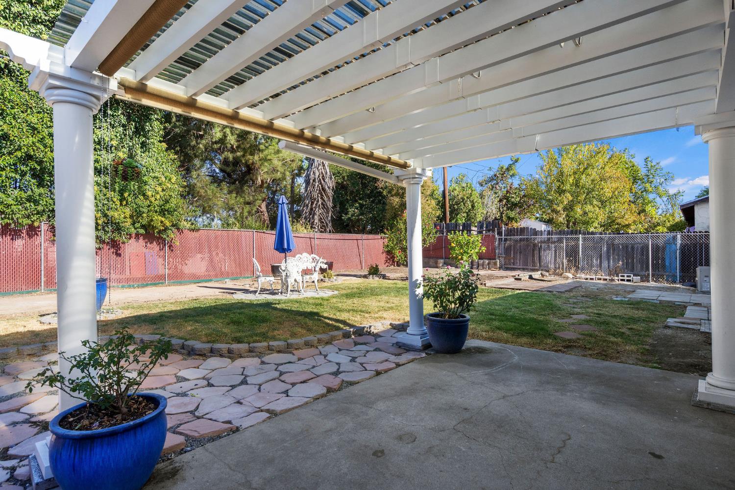 Backyard patio beneath a white pergola, with stone pavers, lawn, and outdoor table and chairs.
