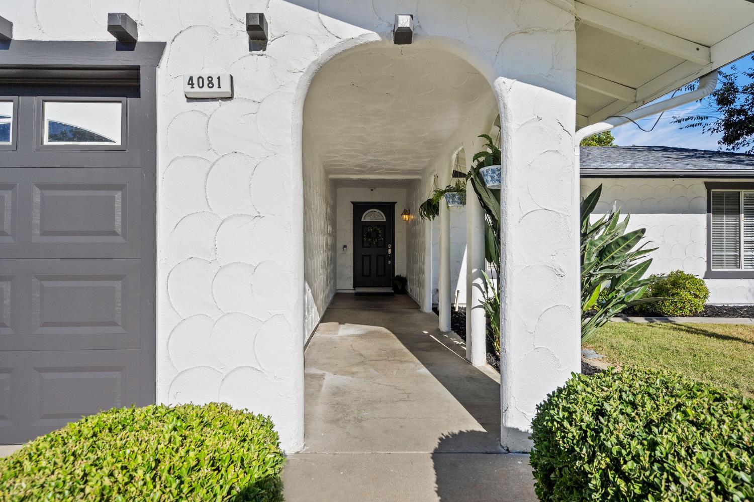 Home entrance with a white stucco arch, covered walkway, and trimmed shrubs.