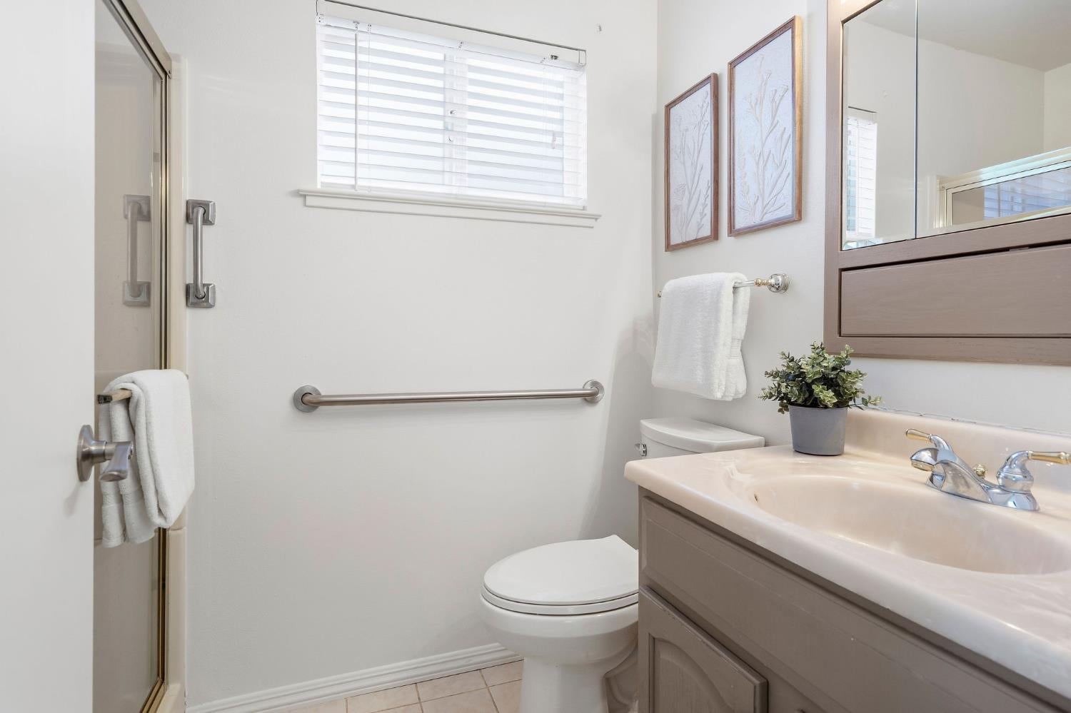 Bathroom with glass shower, beige vanity, toilet, and grab bar beneath a window with white blinds.