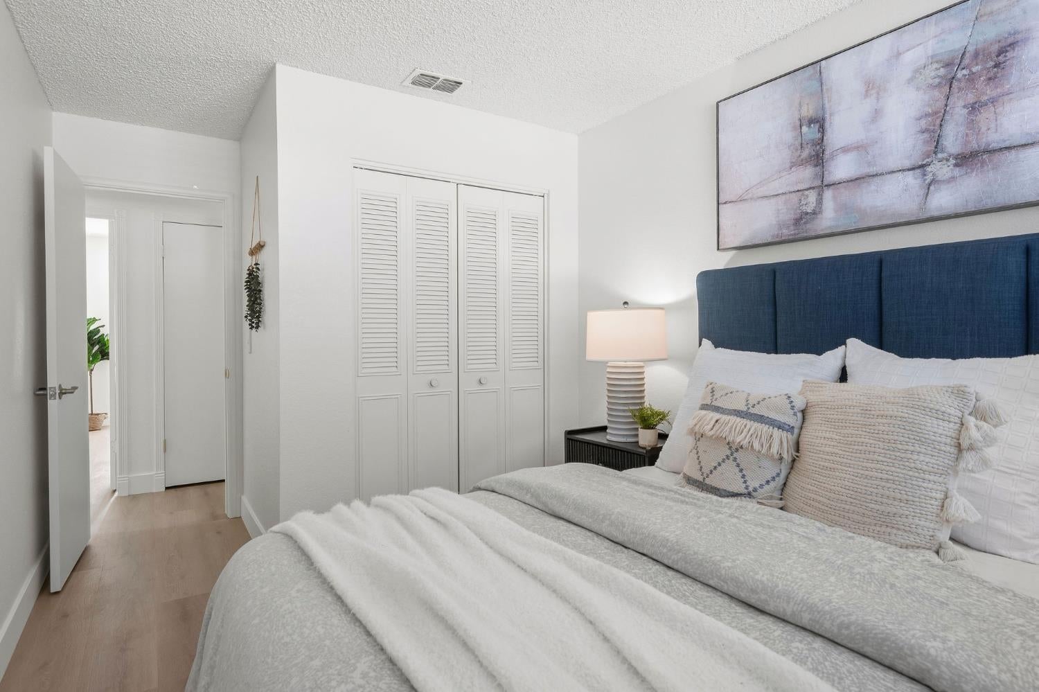 Staged bedroom with a navy headboard, white bedding, louvered closet doors, and abstract wall art.