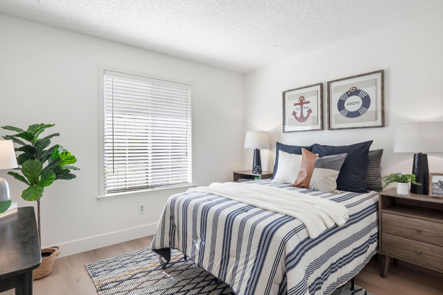 Staged bedroom with striped bedding, nautical wall art, and a large window with white blinds.