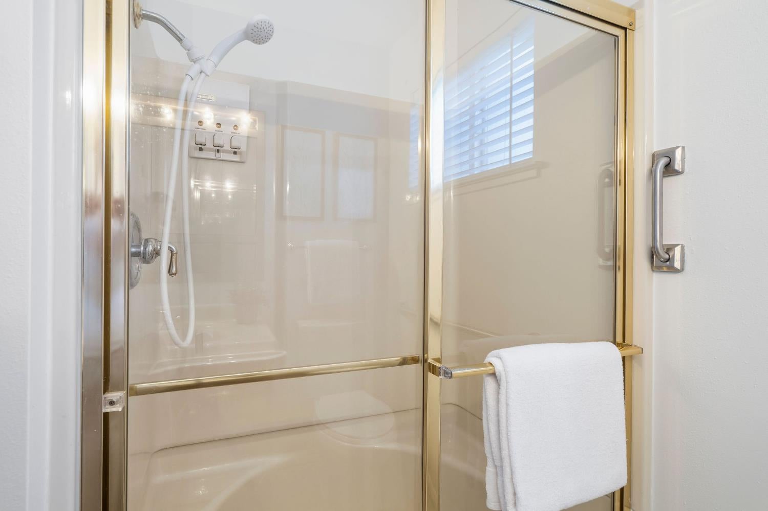 Bathroom shower with gold-tone glass enclosure, handheld showerhead, grab bar, and white towel.
