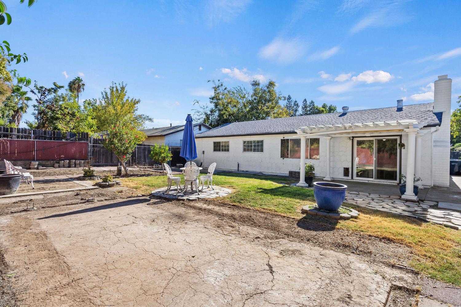 Rear of a white single-story home with a pergola-covered patio, outdoor dining set, and cracked paved yard.