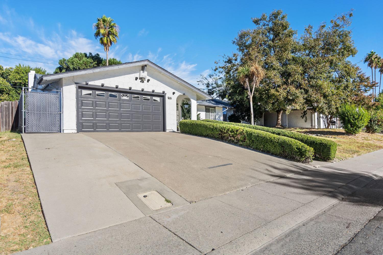 Single-story white house with a dark double garage door, broad driveway, and trimmed hedges.
