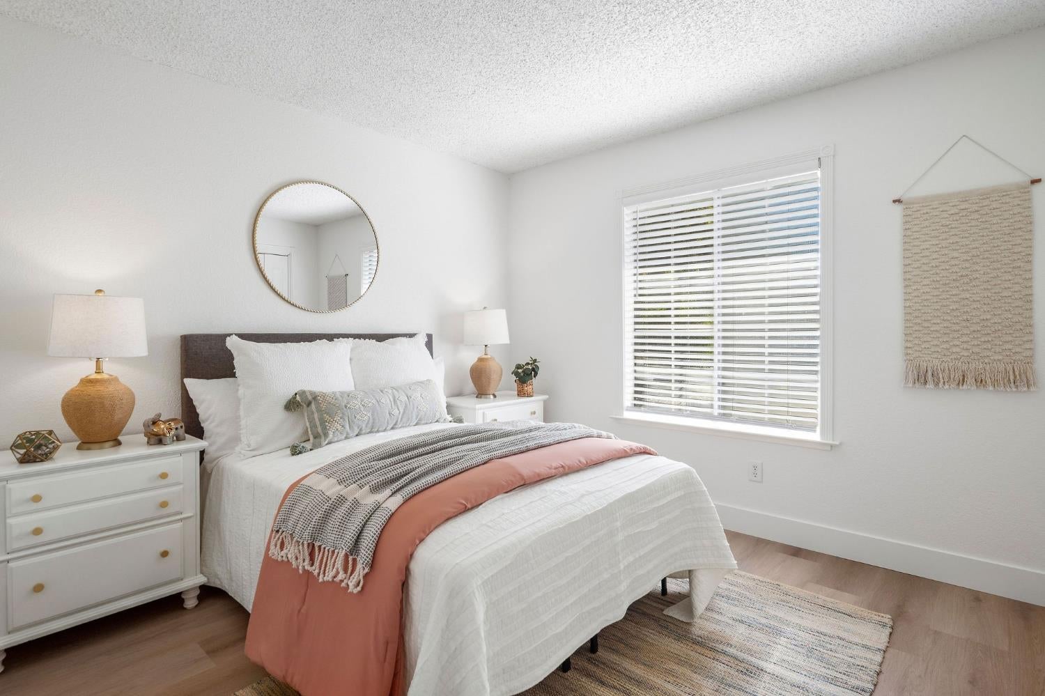 Staged bedroom with a white bed, blush pink throw, round mirror, and window blinds.