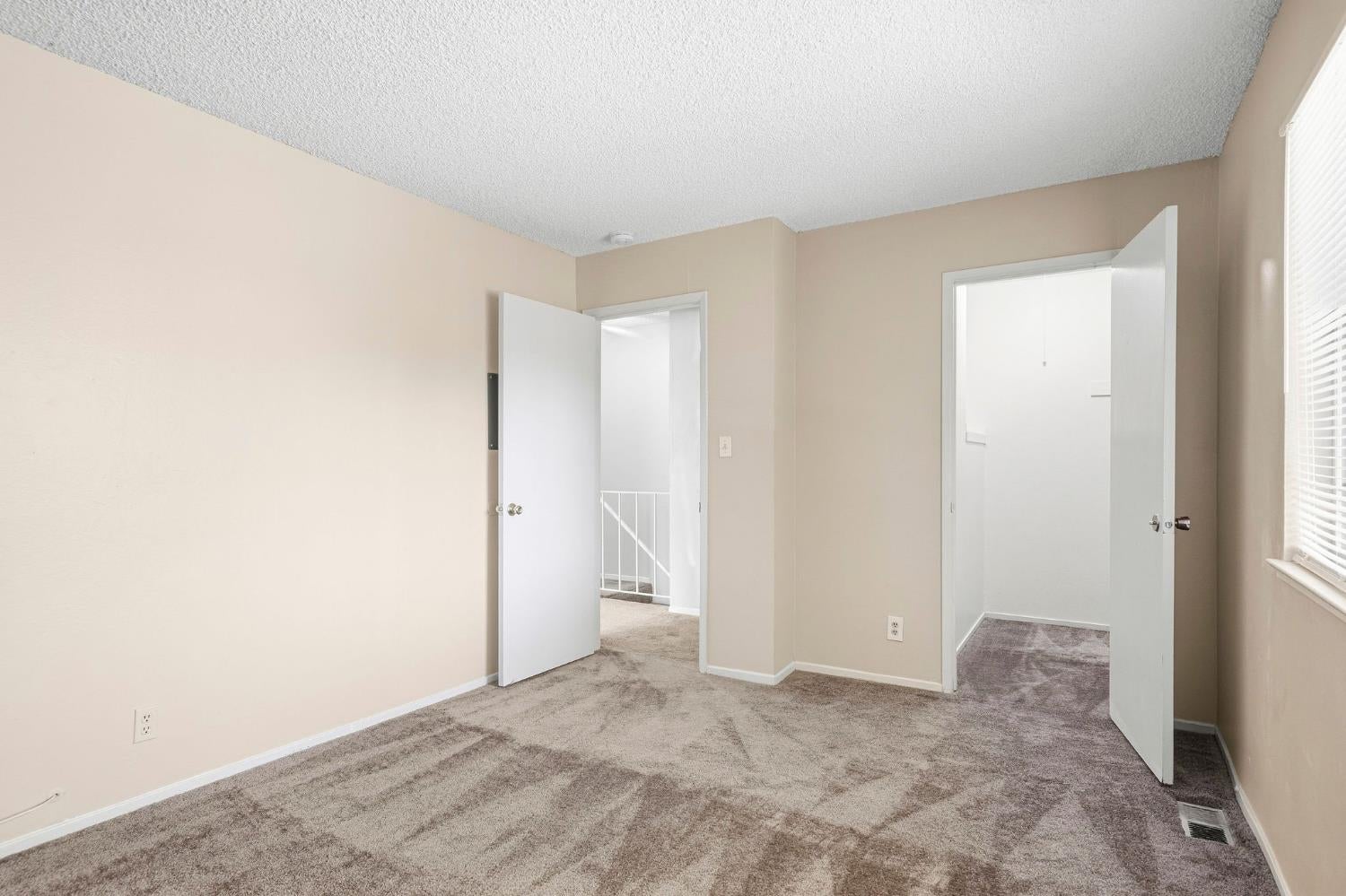 Empty carpeted room with beige walls, a window with blinds, and two open doorways.