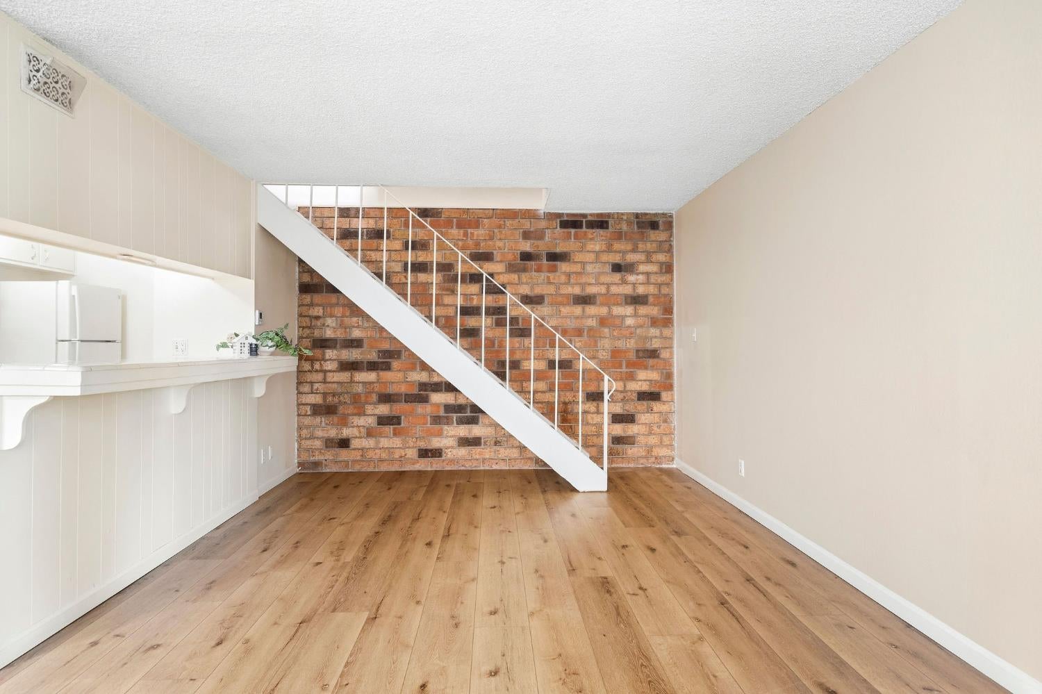Mostly empty interior with wood floors, a white breakfast bar, and a staircase against an exposed brick wall.