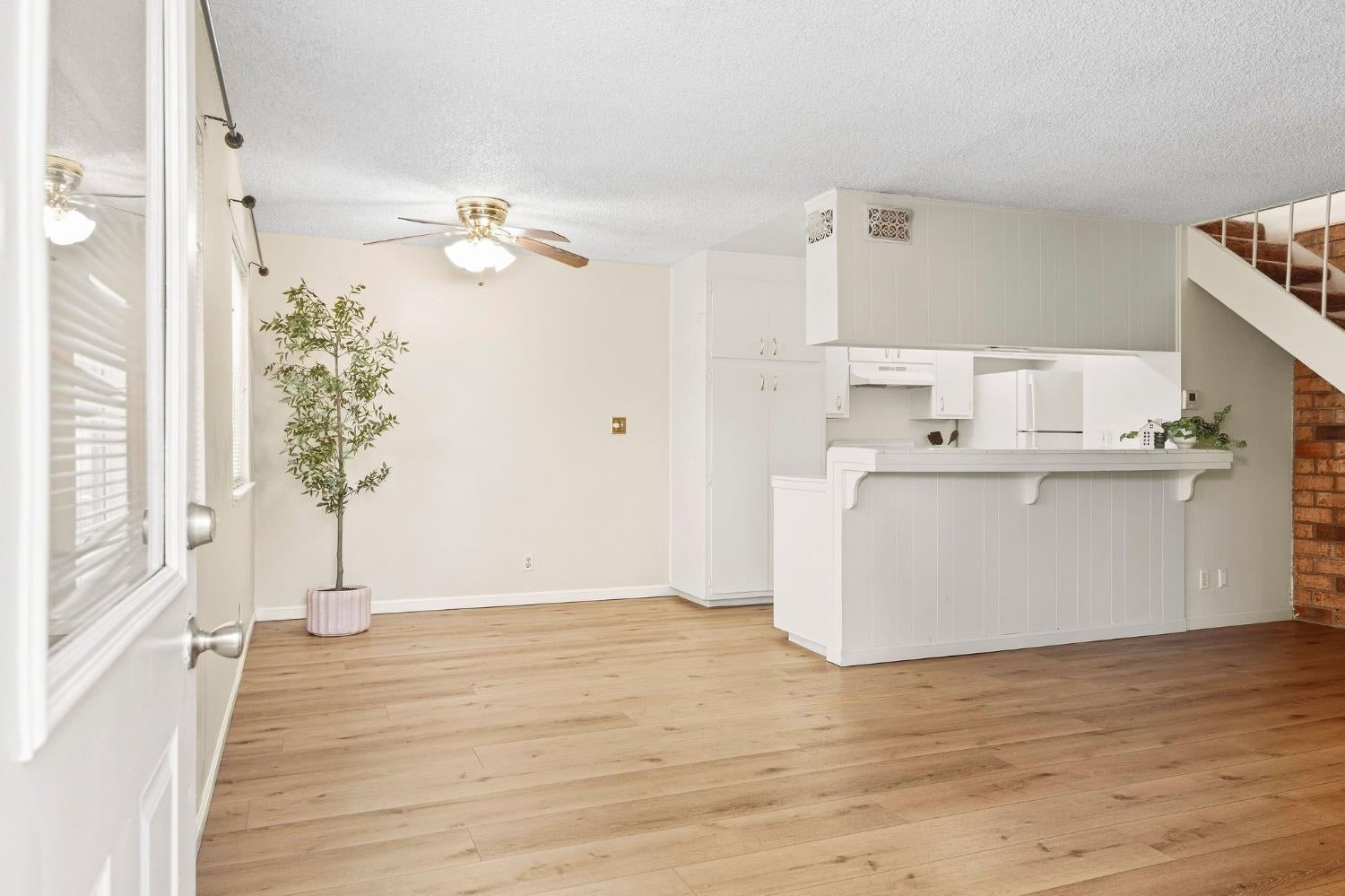 Open living area with light wood floors, a white kitchen peninsula, and a potted tree by the window.