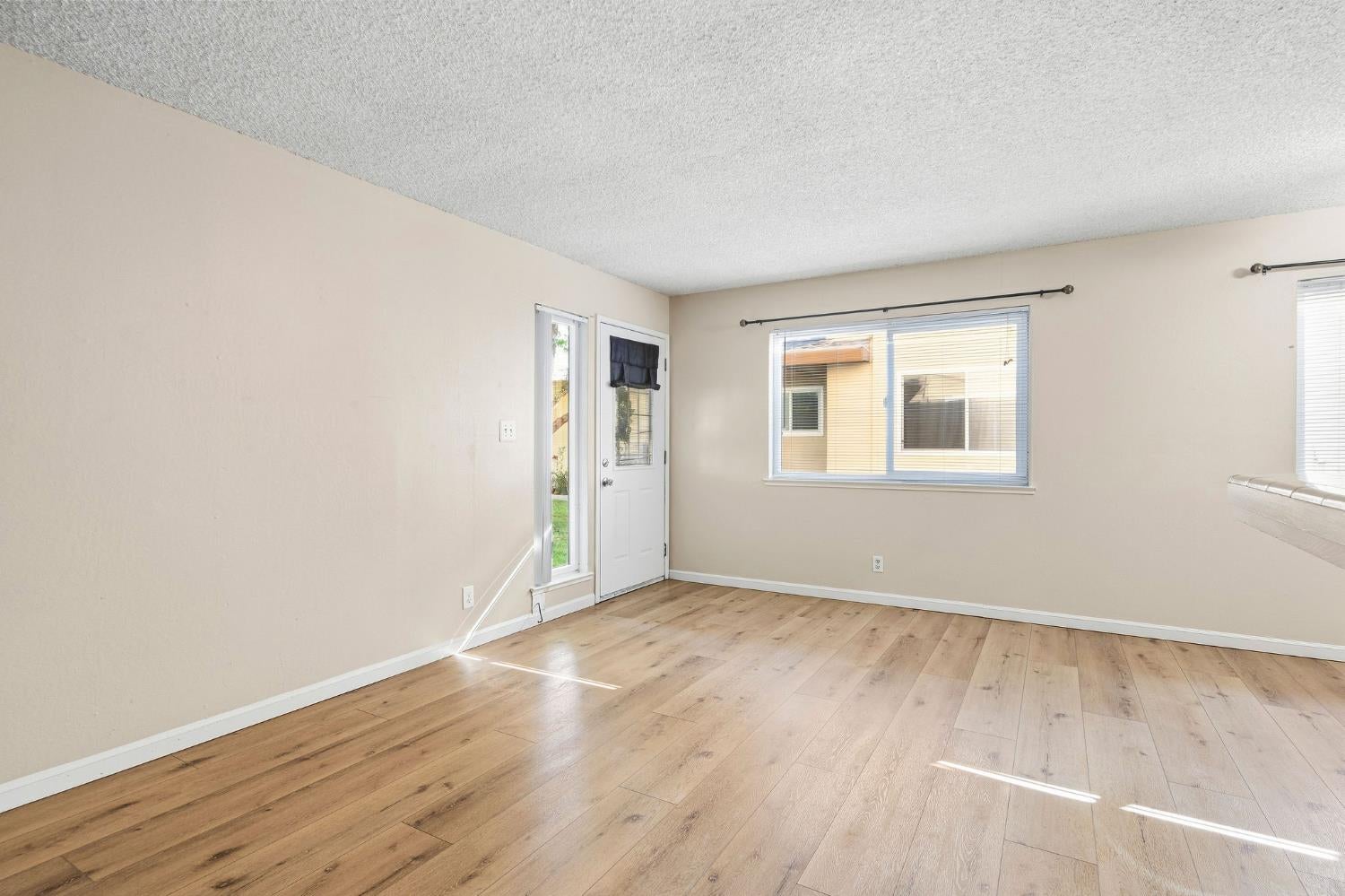 Vacant living room with light wood flooring, beige walls, and a white entry door with sidelight.