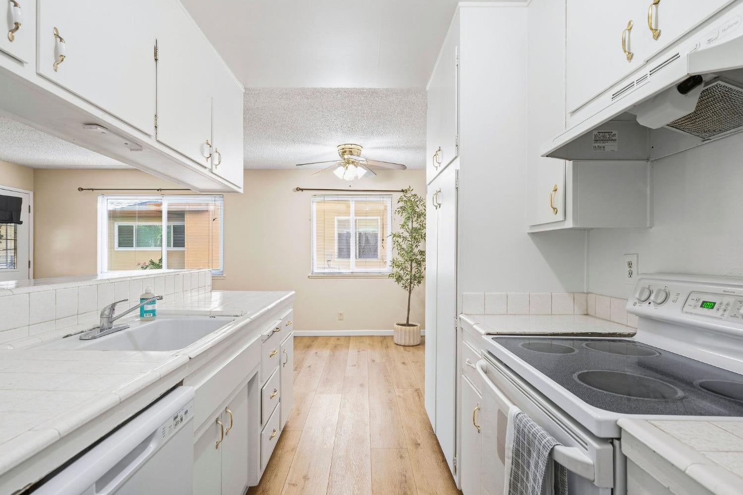 Galley kitchen with white cabinets and tile counters, opening to a dining area with wood floors and a ceiling fan.