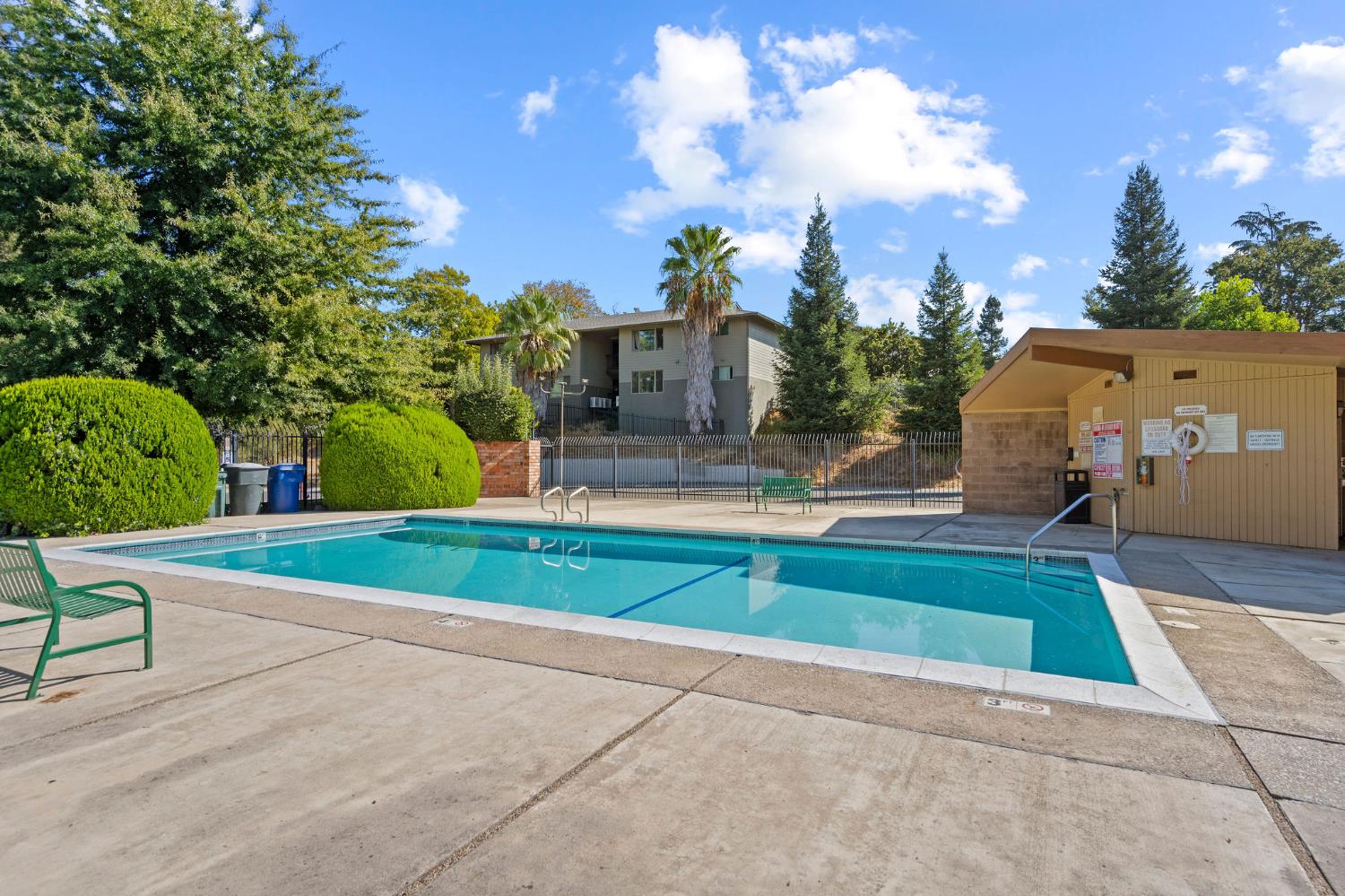 Outdoor community pool beside a bathhouse, with mature trees and a residential building behind it.
