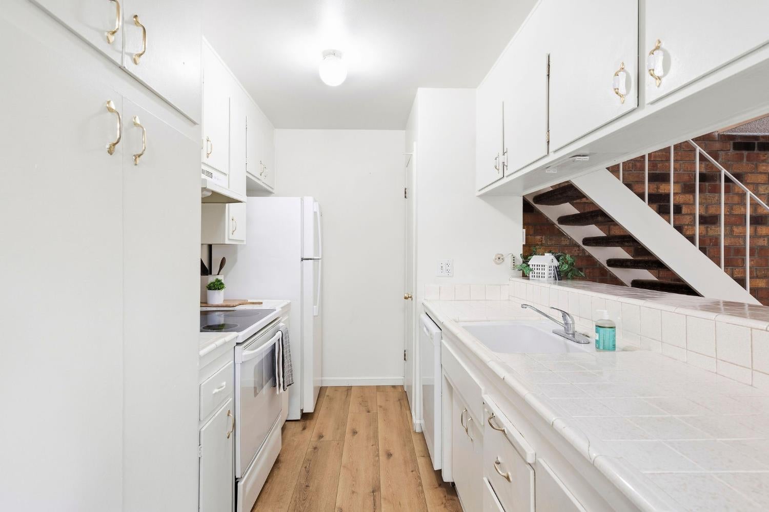 Galley kitchen with white cabinets, tiled counters, wood-look flooring, and a staircase beside a brick wall.
