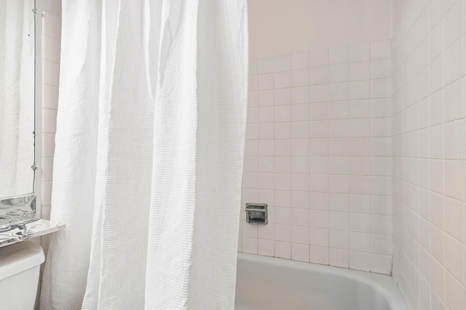 Bathroom with a white bathtub, textured shower curtain, and pale square tile walls.
