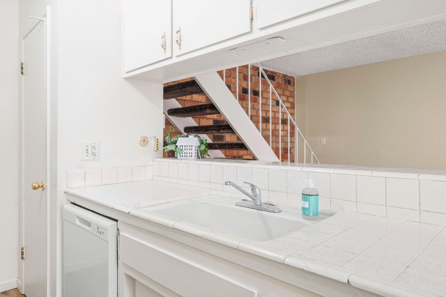Kitchen sink and tiled counter with a dishwasher; brick-backed stairs rise behind it.