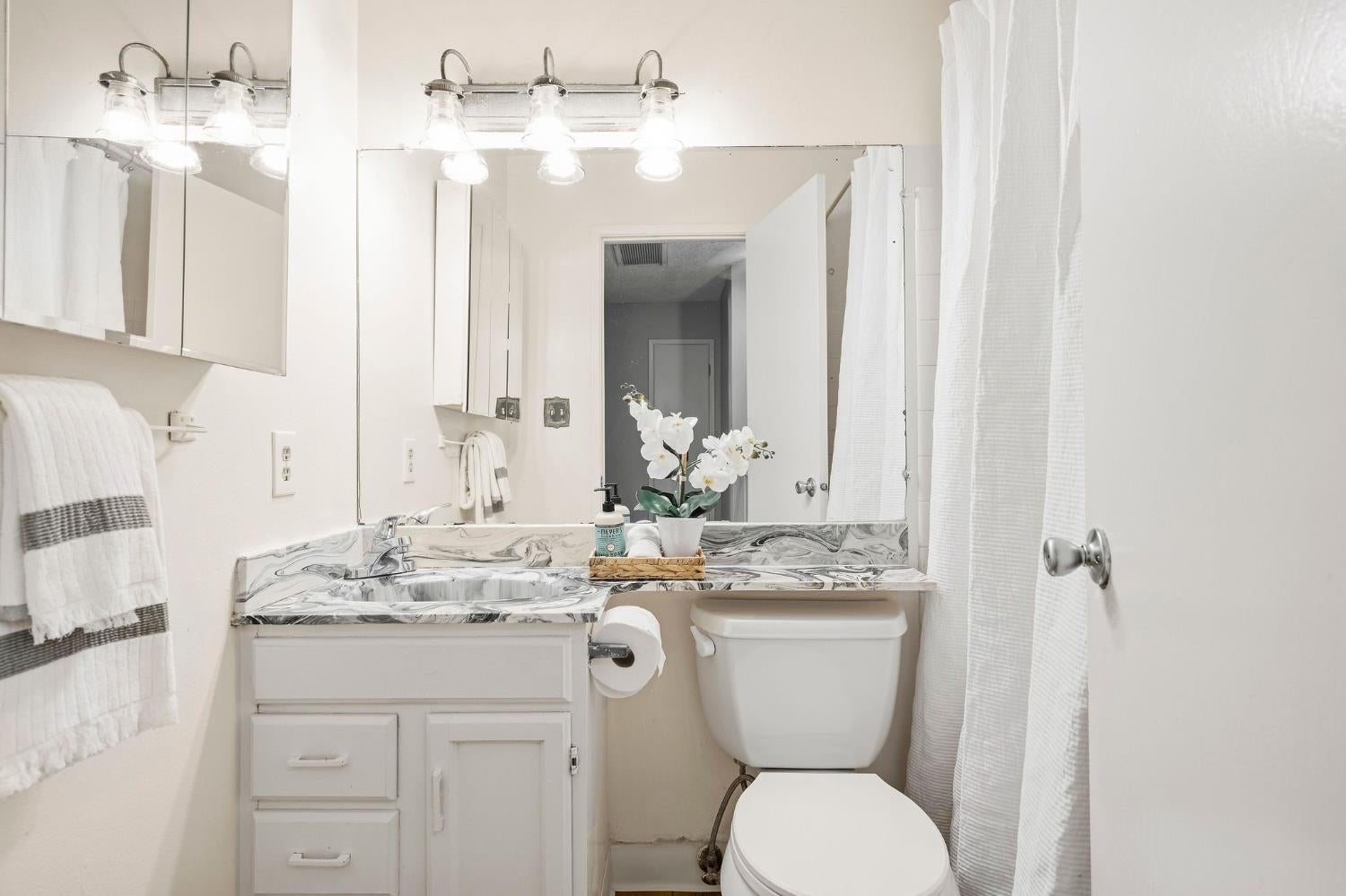 Bathroom with a marble-pattern vanity, mirrored cabinet, toilet, and white shower curtain.