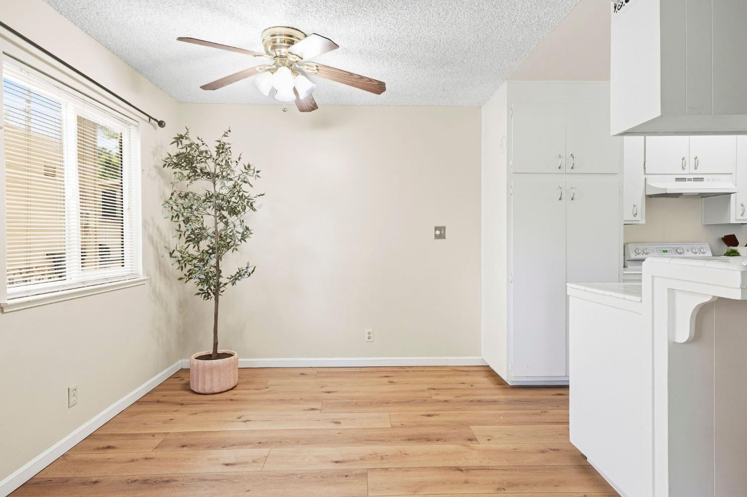 Dining area beside a white kitchen, with wood-look floors, a ceiling fan, and a large window.