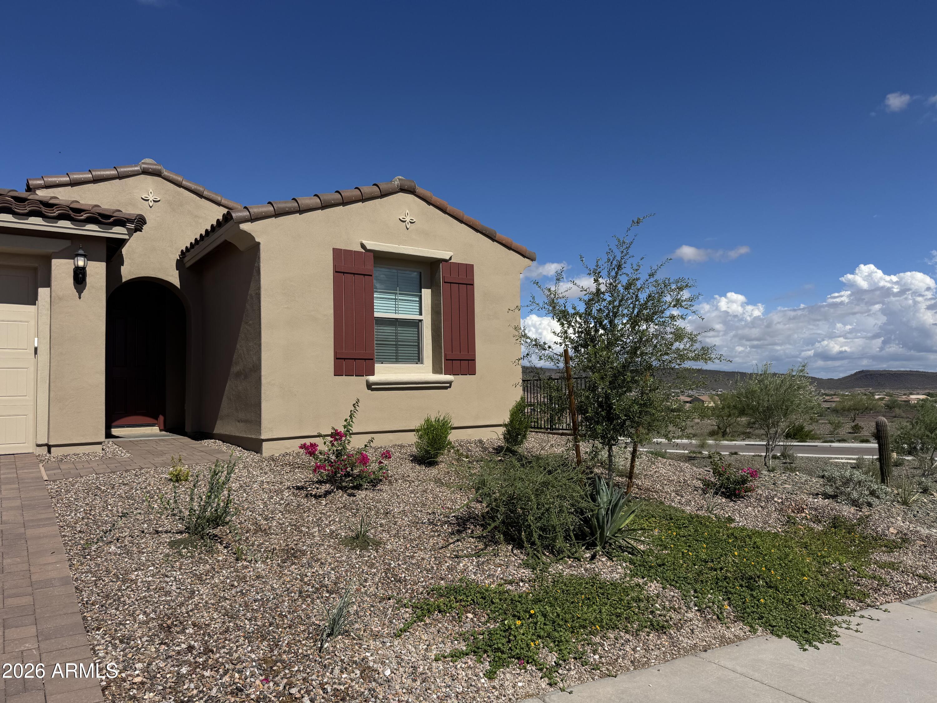 Tan stucco home with red shutters, an arched entry, and desert landscaping beneath a blue sky.