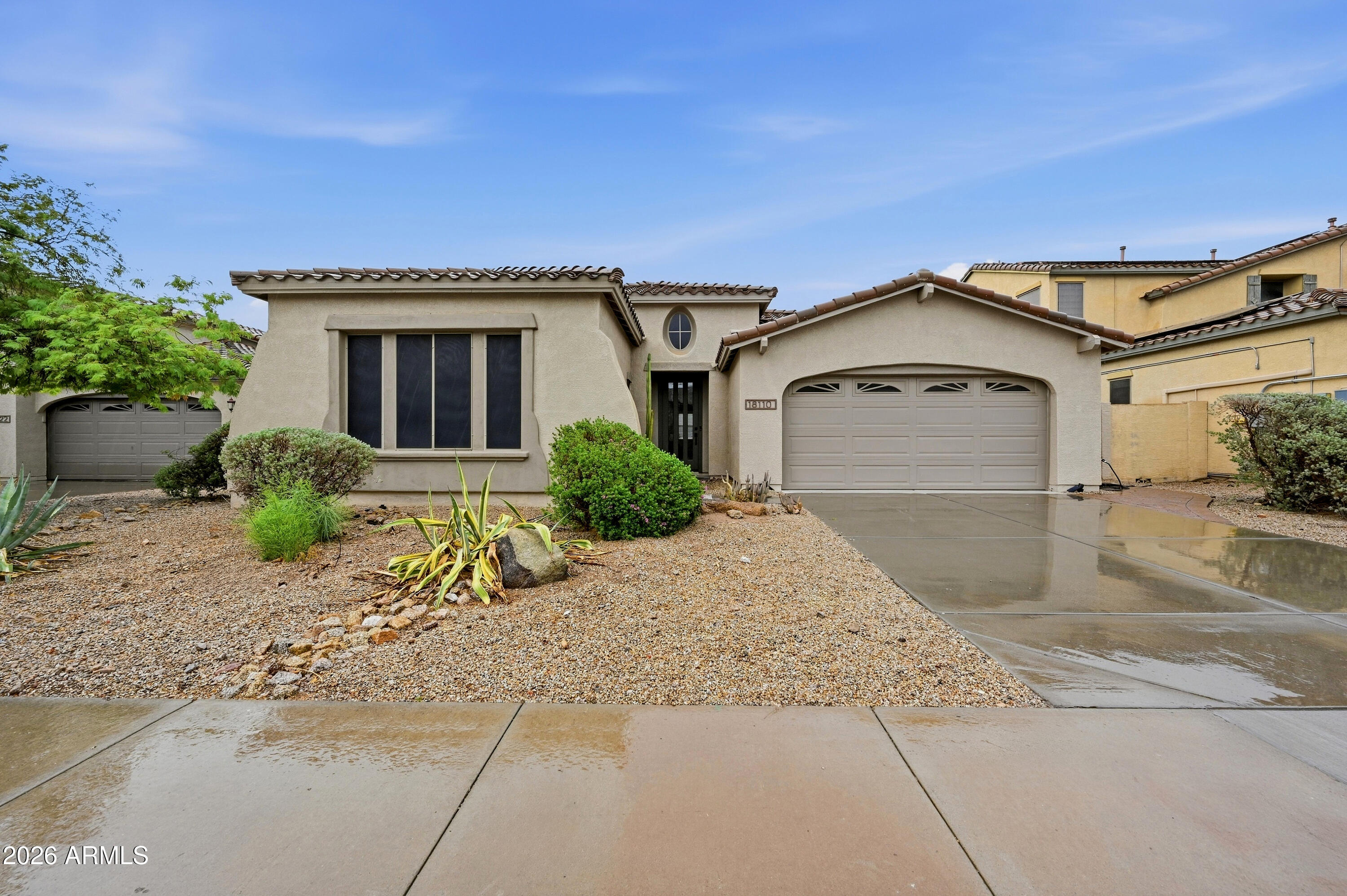 Beige stucco home with a tiled roof, arched garage, gravel landscaping, and wet driveway.