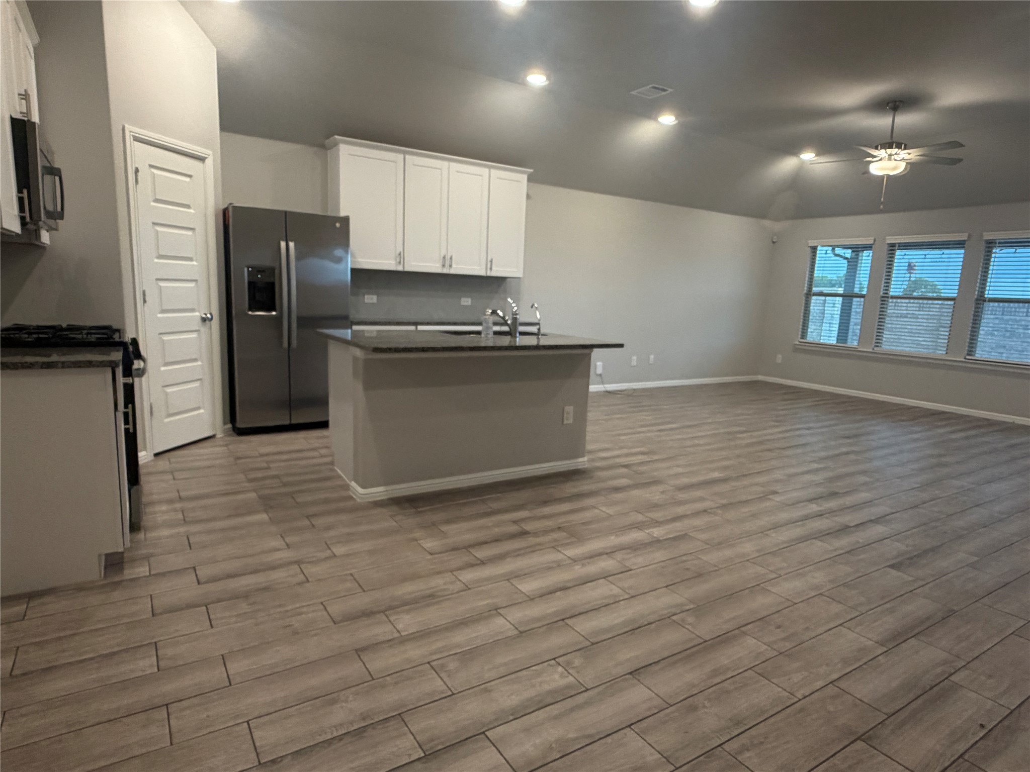 Open-plan kitchen and living area with a central island, stainless steel refrigerator, and wood-look tile floors.