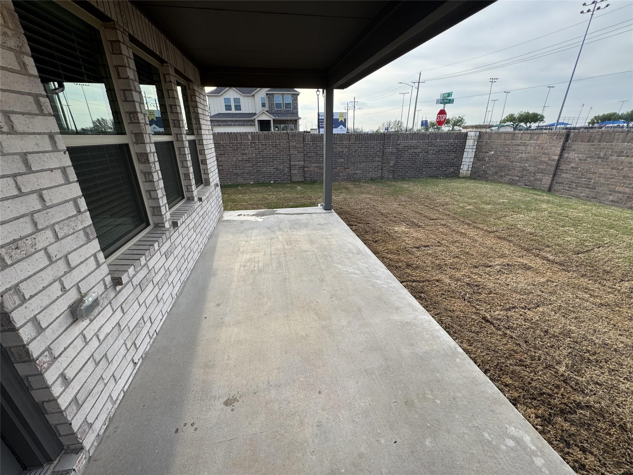 Covered concrete patio beside a brick house, overlooking a fenced yard with sparse grass.