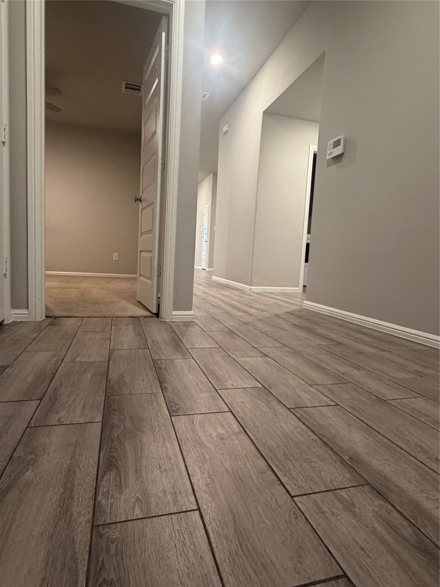 Empty hallway with wood-look tile flooring and an open doorway to a carpeted room.