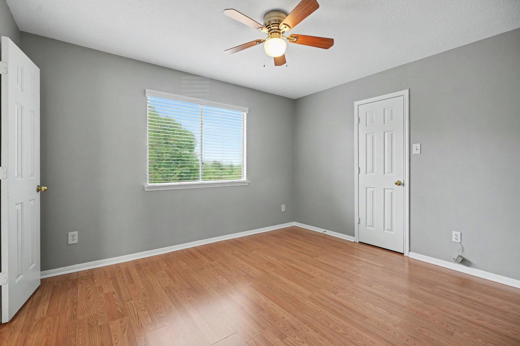Empty room with gray walls, wood flooring, a ceiling fan, and a window overlooking greenery.
