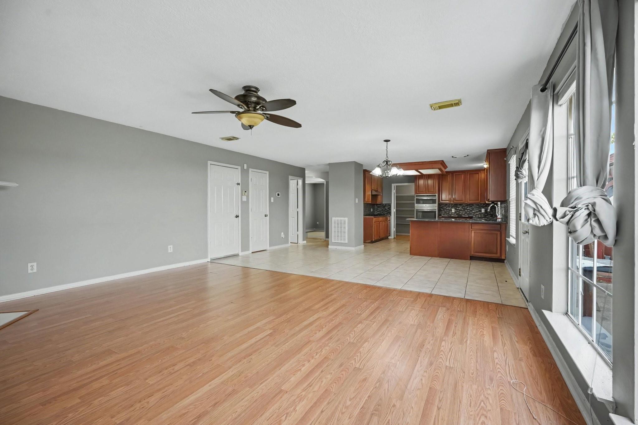 Open living area with wood-look floors, large curtained windows, and a kitchen with reddish-brown cabinets.