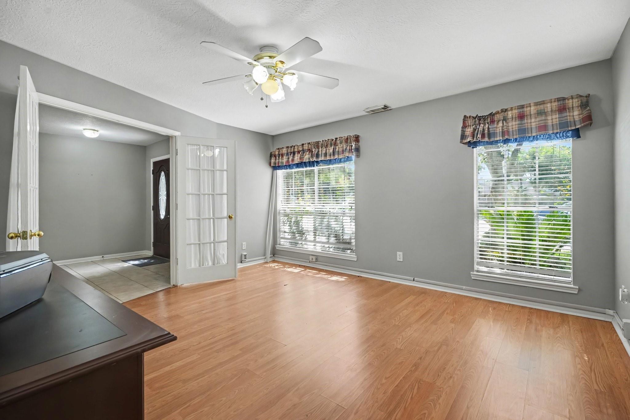 Empty room with wood flooring, two curtained windows, and a ceiling fan beside a tiled entryway.