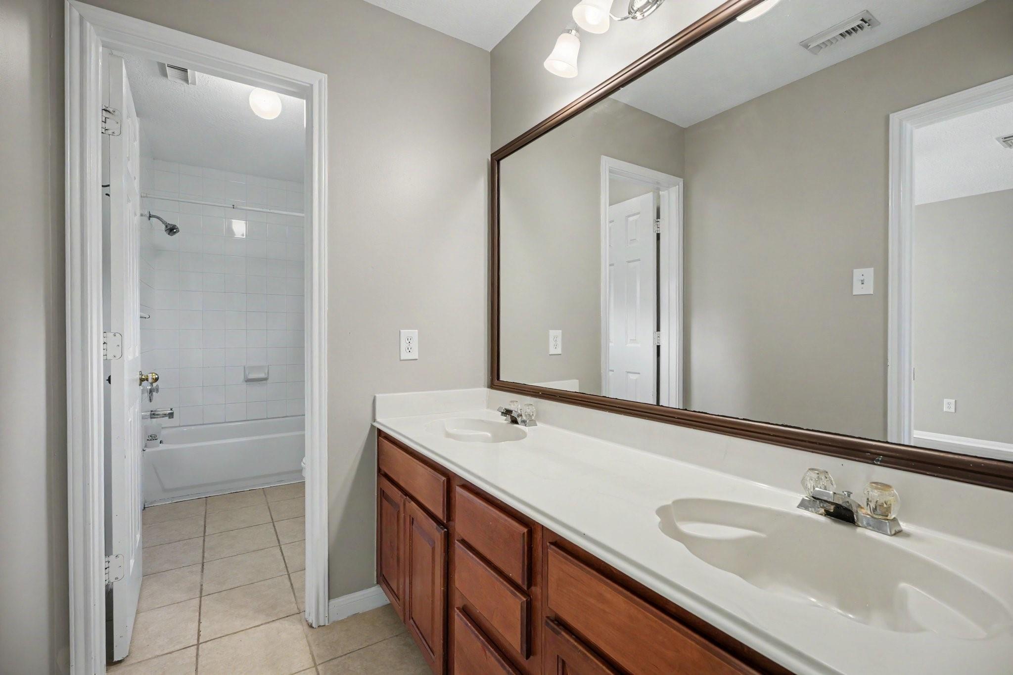 Bathroom with a double-sink vanity and wood cabinets, looking toward a tiled tub and shower.