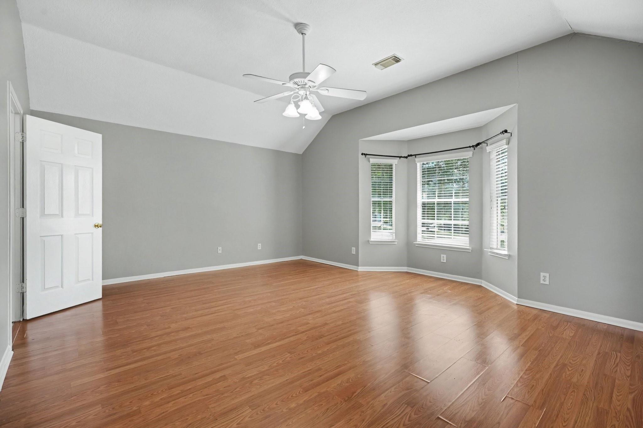 Empty bedroom with vaulted ceiling, wood flooring, and a bay of three windows with blinds.