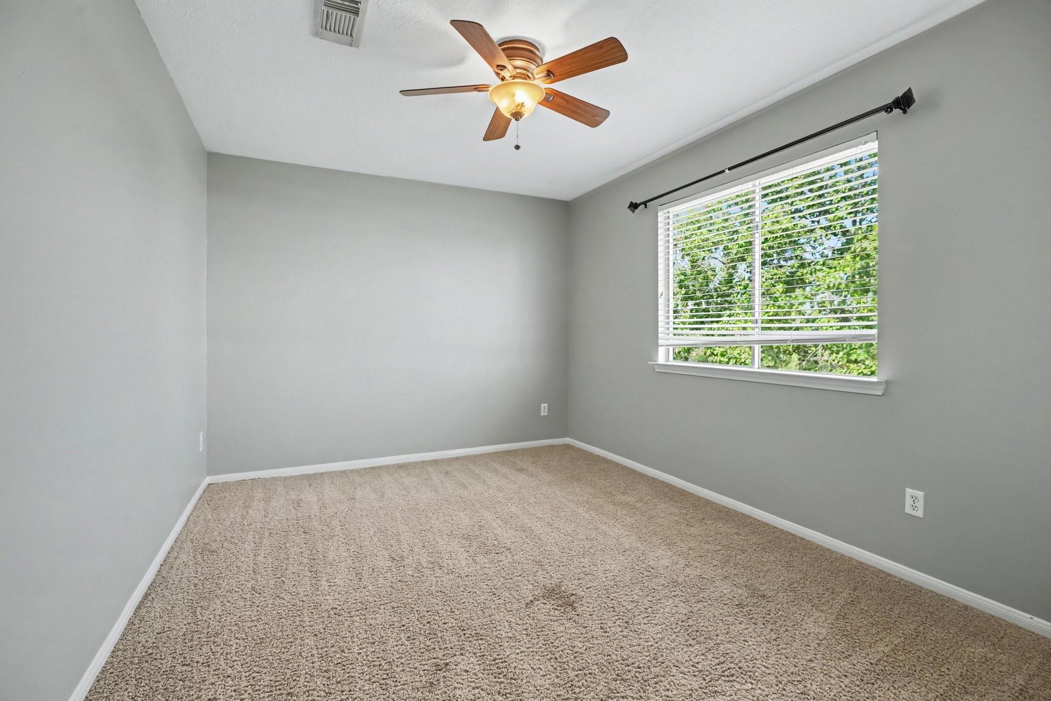 Empty bedroom with beige carpet, a wood-blade ceiling fan, and a window overlooking greenery.