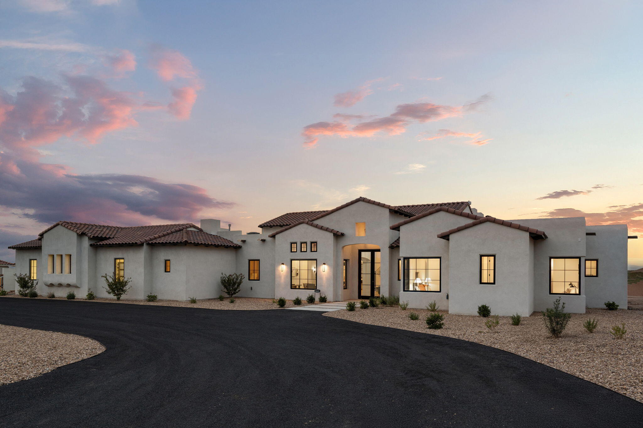 Southwestern stucco home with tiled roofs and glowing windows beside a curved drive at dusk.