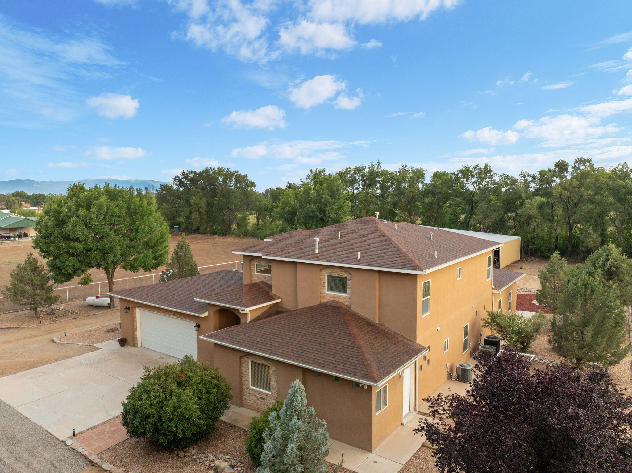 Two-story tan stucco house with a brown roof, broad driveway, mature trees, and distant mountains.